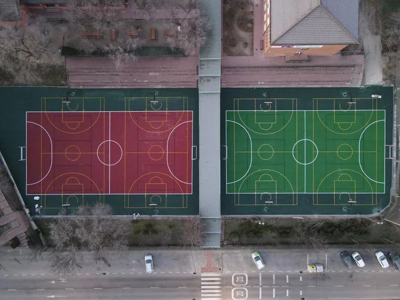 Instalación deportiva en Las Rozas: pistas polideportivas modernas. Vista aérea de dos pistas polideportivas en Las Rozas, una con superficie roja y otra verde, separadas por una pasarela elevada.